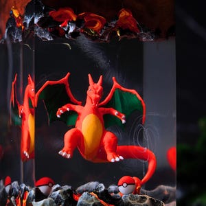 Puede incluir: Escultura de resina con una figura roja de Charizard con alas verdes y vientre amarillo, sobre un fondo oscuro. La escultura incluye dos Poké Balls blancas y rojas y una base similar a la lava, todo encerrado en un bloque rectangular transparente.