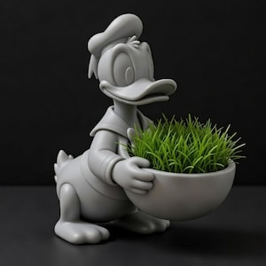 May include: A gray Donald Duck figurine holding a gray bowl filled with green grass. The figurine is in a classic pose, wearing a sailor suit and hat. The background is a solid black.