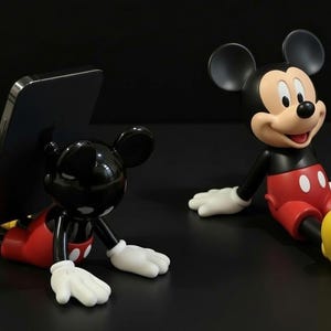 Könnte beinhalten: Zwei Mickey-Mouse-Figuren. Eine hält ein schwarzes Smartphone, die andere sitzt. Beide haben schwarze Ohren, schwarze Körper, rote Shorts, gelbe Schuhe und weiße Handschuhe. Die Figuren stehen auf einer schwarzen Oberfläche.