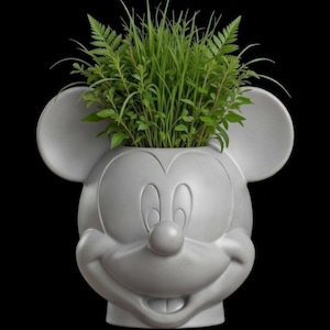 Puede incluir: Una maceta gris con la forma de la cabeza de Mickey Mouse, llena de plantas verdes vibrantes. La maceta tiene una cara sonriente, orejas grandes y un acabado liso. El fondo es negro.