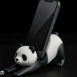 May include: A black smartphone rests in a panda-shaped phone holder. The panda is white with black markings and outstretched arms. The phone holder is made of a glossy material and sits against a black background.