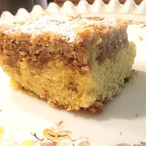 May include: A square slice of yellow crumb cake featuring a thick, textured brown cinnamon streusel topping dusted with white powdered sugar. The cake has a dense, golden-yellow sponge base and is presented on a white plate with floral patterns.