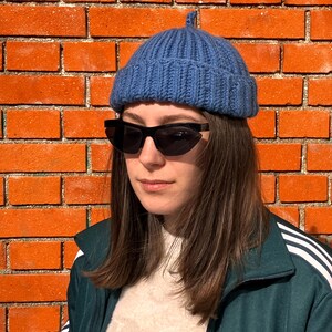 Peut inclure: Un bonnet bleu en tricot avec un petit détail arrondi sur le dessus. Le bonnet est porté avec des lunettes de soleil noires et une veste de survêtement vert foncé avec des rayures blanches et le logo Adidas. En arrière-plan, un mur de briques rouges.