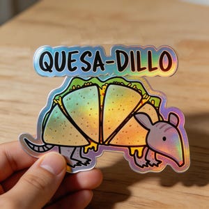 May include: Holographic sticker featuring a cartoon armadillo with a quesadilla body. The sticker has the text "Quesad-dillo" in a bubble-style font. The armadillo has a light purple head and feet, with a yellow and green quesadilla body.