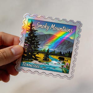 May include: A holographic sticker featuring a Smoky Mountains landscape with a rainbow, river, and bear. The sticker has a postage stamp design with the text "Smoky Mountains" and "Tennessee".
