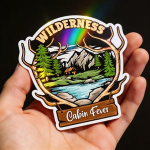 May include: A sticker with a wilderness scene, featuring a rainbow, mountains, trees, and a cabin. The word "WILDERNESS" arches across the top, and "Cabin Fever" is written on a wooden sign at the bottom. The sticker has a gold border.