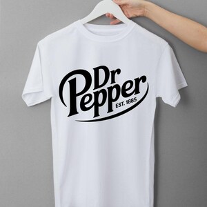 May include: White t-shirt with the Dr Pepper logo in black. The logo includes the words "Dr Pepper" in a stylized font, with "EST. 1885" below. The shirt is displayed on a white hanger.