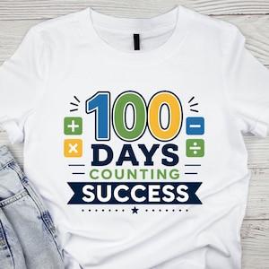 Può includere: T-shirt bianca con una grafica colorata che celebra i 100 giorni di scuola. Il design presenta il numero "100" in blu, verde e giallo, insieme alle parole "Days Counting Success". Include simboli matematici.