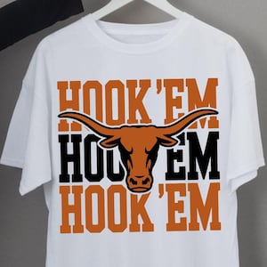 May include: A white short-sleeved t-shirt featuring a graphic print with the text HOOK 'EM in orange and black block lettering. A central illustration of a longhorn bull head in orange is positioned between the lines of text on the shirt.