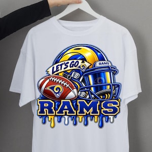 Pode incluir: Uma t-shirt branca de manga curta com um gráfico de um capacete de futebol americano azul e amarelo, uma bola de futebol americano castanha e o texto "LET'S GO" e "RAMS" em azul e amarelo. O design inclui um efeito de tinta a escorrer.