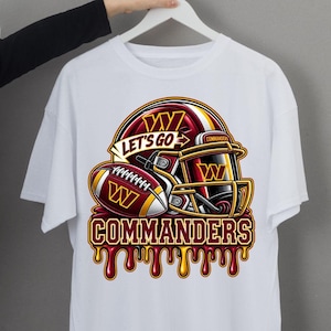 Pode incluir: Uma t-shirt branca de manga curta com um gráfico de um capacete de futebol americano e uma bola nas cores bordô e dourado. O design inclui o texto "LET'S GO" e "COMMANDERS" em fonte negrito, com um efeito de tinta a escorrer abaixo.