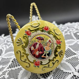May include: A round, pale yellow metal tin featuring a central circular illustration of two figures in 18th-century attire. The lid is decorated with gold-toned scrollwork and pink rose accents. The tin is supported by a gold-toned twisted metal stand.