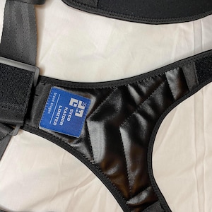 May include: A black harness featuring adjustable nylon straps and a padded, quilted back panel. A blue rectangular label is stitched onto the center, displaying the white text SYED HAIDER LIMITED and the letters SH in a square logo design.
