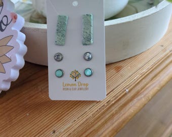 May include: A set of earrings on a white card. The earrings include two pairs of stud earrings and a pair of rectangular earrings. The earrings are light green with a speckled design. The card has the text "Lemon Drop Resin & Clay Jewellery".