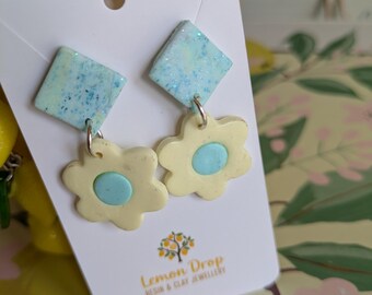 May include: A pair of handmade earrings featuring a light yellow flower design with a blue center, suspended from a blue square. The earrings are attached to a white card with the text "Lemon Drop Resin & Clay Jewellery".