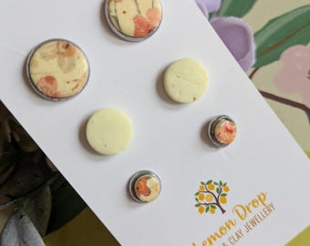 May include: A set of handmade stud earrings on a white card. The earrings feature a floral design with orange and yellow hues, set in silver-colored metal. The card has the logo "Lemon Drop Resin & Clay Jewellery".