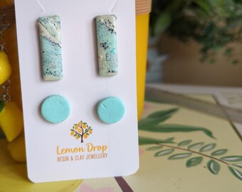 May include: A pair of earrings on a white card. The earrings include two rectangular studs with blue and white marbling and two round, light blue studs. The card has the text "Lemon Drop Resin & Clay Jewellery".