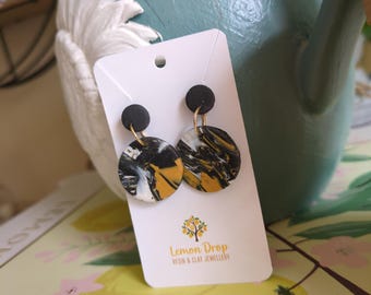 May include: A pair of handmade earrings with a black, white, and gold marbled design. The earrings are attached to a white card with the "Lemon Drop Resin & Clay Jewellery" logo. The earrings have a black stud and gold-colored hardware.