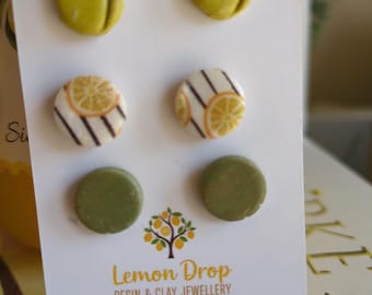 May include: A set of six stud earrings in various designs. Two are olive green tulip shapes, two are white with orange citrus slices and black stripes, and two are olive green circles. The card reads "Lemon Drop Resin & Clay Jewellery."