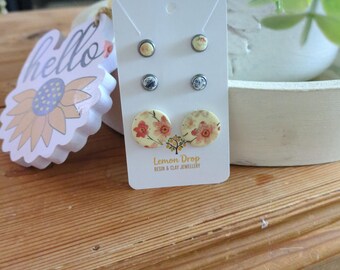 May include: A set of three pairs of stud earrings on a white card. The earrings feature floral designs in yellow, red, and blue. The card has the text "Lemon Drop Resin & Clay Jewellery". A wooden surface is in the foreground.