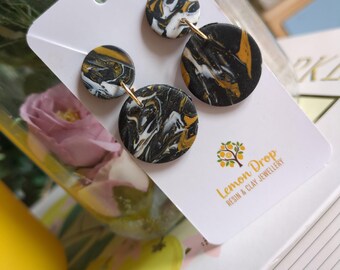 May include: A pair of handmade earrings featuring a marbled design in black, white, and gold. The earrings are round and connected with gold-colored metal. The earrings are displayed on a white card with the text "Lemon Drop Resin & Clay Jewellery".