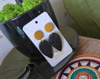 May include: A pair of handmade earrings with a yellow circle top and a black teardrop-shaped bottom with a floral pattern. The earrings are displayed on a white card with the text "Lemon Drop Resin & Clay Jewellery".