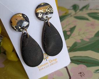 May include: A pair of black teardrop earrings with gold accents and a gold-colored hoop. The top of the earrings are round with black, gold, and white patterns. The earrings are displayed on a white card with the text "Lemon Drop Resin & Clay Jewellery."
