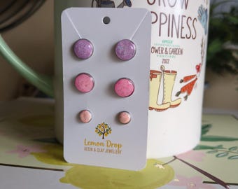 May include: A set of six stud earrings in shades of pink and purple, displayed on a white card. The earrings are resin and clay jewelry. The card has the text "Lemon Drop RESIN & CLAY JEWELLERY". A white mug with the text "GROW HAPPINESS" is in the background.