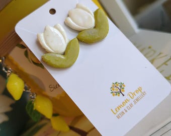 May include: A pair of white and green tulip-shaped earrings on a white card. The card has the text "Lemon Drop Resin & Clay Jewellery" and a lemon tree logo. The earrings are on a yellow background.