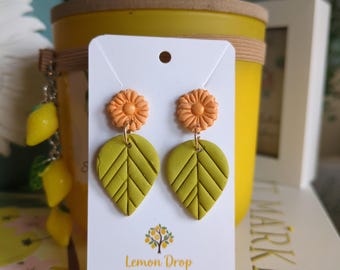 May include: A pair of handmade earrings featuring orange flower studs and olive green leaf-shaped dangles. The earrings are displayed on a white card with the text "Lemon Drop Resin & Clay Jewellery". A yellow candle and lemon-shaped charm are in the background.