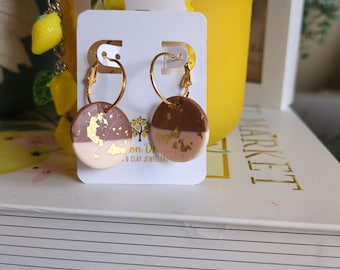 May include: Gold hoop earrings with round, two-tone clay pendants. The pendants are half light pink and half brown, with gold leaf accents. A yellow container and lemon charm are in the background. The earrings are displayed on a white card.