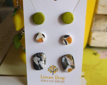 May include: A white card displays three pairs of earrings. The top pair is olive green, the middle pair has a white, black, and orange pattern, and the bottom pair is black, white, and gold. The card reads "Lemon Drop Resin & Clay Jewellery."