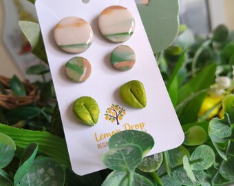 May include: A set of handmade stud earrings on a white card. The earrings feature a variety of designs, including round studs with orange, green, and cream colors, and leaf-shaped studs in a metallic green. The card has the text "Lemon Drop Resin".