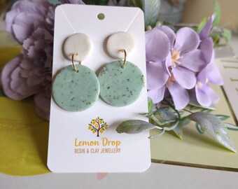 May include: A pair of handmade earrings with a white top circle and a speckled mint green bottom circle. The earrings are attached to a white card with the "Lemon Drop" logo. The background includes purple flowers and greenery.