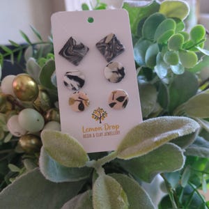May include: A selection of six stud earrings displayed on a white card. The earrings showcase diverse designs: marbled black and white squares, circular black and white patterns, and beige circles with black leaf details. The card is labelled "Lemon Drop Resin & Clay Jewellery."