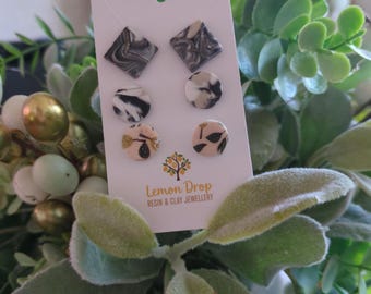 May include: A set of six stud earrings on a white card. The earrings feature various designs: marbled black and white squares, circular black and white patterns, and beige circles with black leaf accents. The card reads "Lemon Drop Resin & Clay Jewellery."