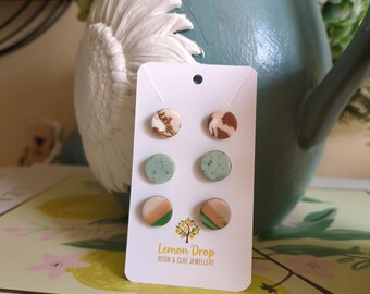 May include: A set of six stud earrings on a white card. The earrings feature various designs, including marbled patterns in brown, green, and orange, and speckled gold accents. The card has the text "Lemon Drop RESIN & CLAY JEWELLERY".