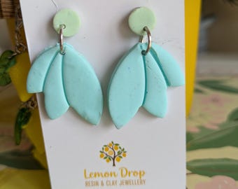May include: A pair of handmade earrings featuring a light blue petal design with a mint green accent. The earrings are attached to a white card with the text "Lemon Drop Resin & Clay Jewellery" and a logo of a tree with yellow fruit.