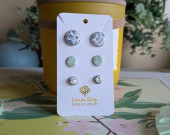 May include: A set of stud earrings on a white card with the text "Lemon Drop Resin & Clay Jewellery." The earrings feature various designs, including blue floral patterns, speckled green, and white with black accents. The background includes a yellow pot and floral decorations.