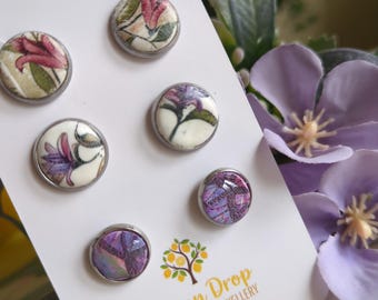 May include: A set of six floral resin and clay jewelry pieces. The round pieces feature various floral designs in shades of pink, purple, and green, set in silver-toned metal. The card reads "Lemon Drop Resin & Clay Jewellery."
