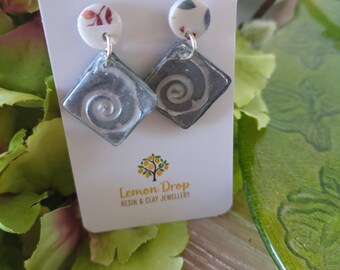 May include: A pair of handmade earrings with a floral design on the top circle and a gray, square pendant with a spiral design. The earrings are displayed on a white card with the "Lemon Drop Resin & Clay Jewellery" logo.