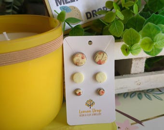 May include: A yellow candle and a card of resin and clay earrings are displayed. The earrings have floral designs and solid yellow circles. The card reads "Lemon Drop Resin & Clay Jewellery." A sign reads "Where troubles melt like drops."