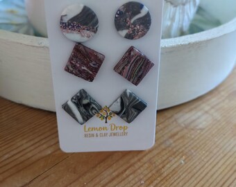 May include: A white card displays six pairs of stud earrings in various shapes. The earrings feature marbled designs in shades of black, white, and burgundy, with some including glitter accents. The card is labeled "Lemon Drop Resin & Clay Jewellery."