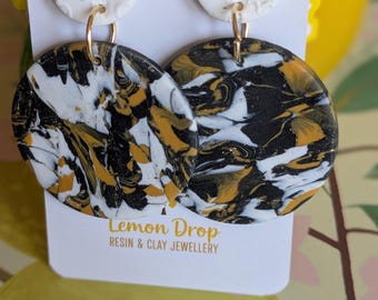 May include: A pair of handmade earrings with a floral design. The earrings feature a white top circle with a flower pattern and a larger, marbled bottom circle in black, white, and gold. The earrings are connected with gold-colored hoops. The tag reads "Lemon Drop Resin & Clay Jewellery."