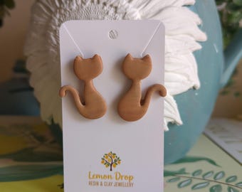 May include: A pair of light brown cat-shaped earrings on a white card with the text "Lemon Drop Resin & Clay Jewellery". The earrings are displayed against a blurred background featuring a blue vase and floral decorations.