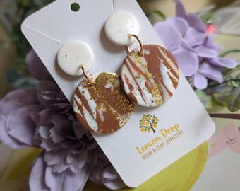 May include: A pair of handmade earrings featuring a white top circle and a larger, round bottom with brown, white, and gold marbling. The earrings hang from gold-colored hoops. The earrings are displayed on a white card with the Lemon Drop logo.