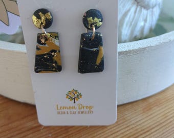 May include: A pair of black and gold earrings with a rectangular shape and gold leaf accents. The earrings are attached to a small, round black stud. The earrings are displayed on a white card with the text "Lemon Drop Resin & Clay Jewellery".