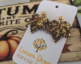 May include: A pair of dark brown maple leaf-shaped earrings with gold leaf accents and gold-tone ball studs, attached to a white display card featuring the text Lemon Drop RESIN & CLAY JEWELLERY and a small tree graphic with yellow lemon-like fruits.