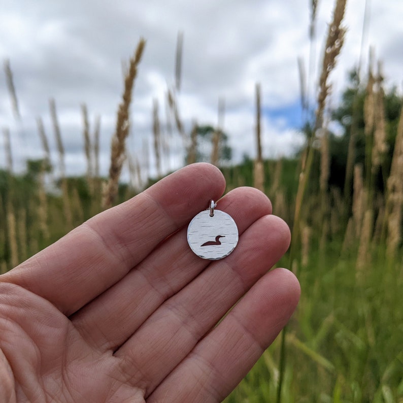 May include: A silver pendant featuring a cut-out loon design. The pendant is round and has a textured surface. The loon is facing to the right.