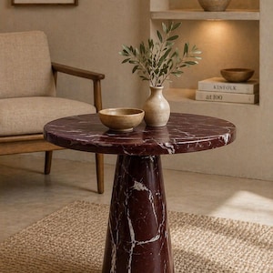 May include: A round, burgundy marble side table with white veining. The table has a single, conical base. A small vase with greenery and a wooden bowl sit on top. The table is on a woven rug.